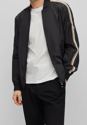 Mann trägt schwarze Bomberjacke mit Reißverschluss und beige-weißen Streifen an den Ärmeln, weißes T-Shirt und schwarze Hose, Hände seitlich am Körper.