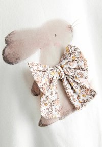 Weißer Stoff mit einem aufgedruckten rosa Hasen mit brauner Nase, verziert mit einer Schleife im floralen Muster in sanften Beige-, Gelb- und Brauntönen.