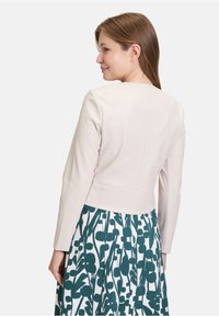 Femme portant une veste courte beige clair et une jupe blanche avec un motif de feuilles abstrait vert foncé, vue de dos et légèrement tournée sur le côté.