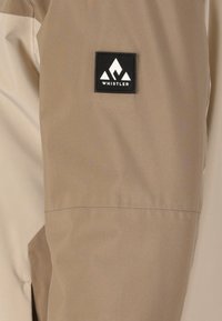 Tan jakke stof med en glat tekstur, der har et sort firkantet logo-patch med teksten "WHISTLER" og bjergikoner.