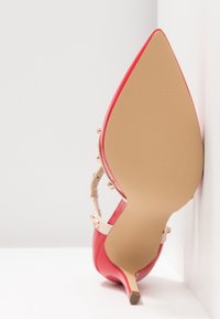 Chaussure à talon haut rouge à bout pointu et semelle beige, reposant droite sur le talon contre un mur blanc d’angle.