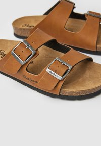 Sandales en cuir marron avec un semelle en liège, deux brides ajustables avec des boucles en argent, et une semelle en caoutchouc noire texturée.