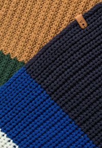 Couverture tricotée avec des sections en marron, vert, bleu et marine, présentant un motif texturé tissé et une petite étiquette en cuir.