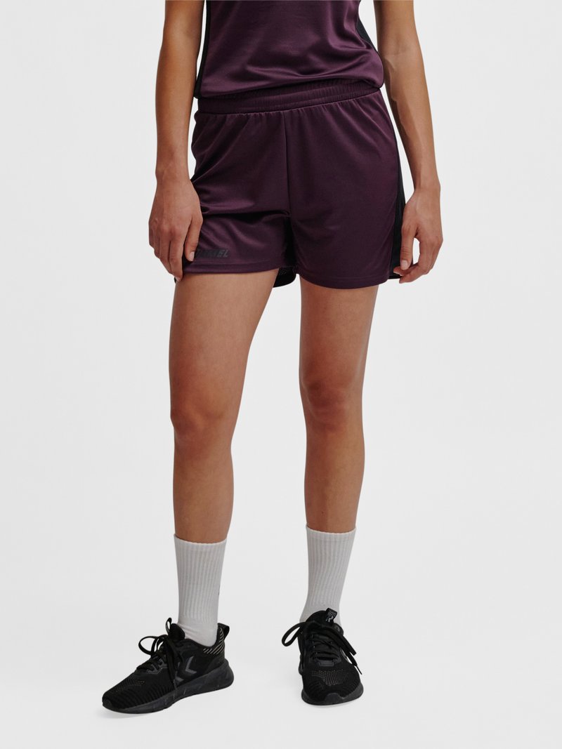 Persoon draagt donkerpaarse sportshorts, witte enkelsokken en zwarte hardloopschoenen, staand tegen een effen lichte achtergrond.