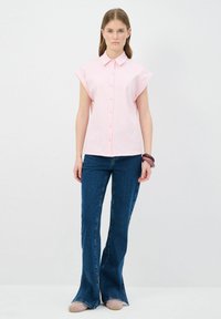 Camicia rosa chiaro a maniche corte con bottoni abbinata a jeans di denim blu a zampa d'elefante. La camicia presenta un colletto e una consistenza sottile.