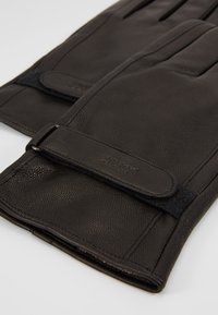 Schwarze Lederhandschuhe mit strukturierter Oberfläche, die eng anliegen und über einen Stoffbandverschluss mit geprägt aufgedrucktem Logo am Riemen verfügen.