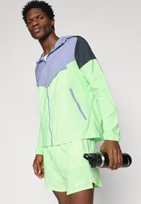 Homme portant une veste de sport néon vert et gris clair, ainsi que des shorts, tenant une bouteille d'eau Nike noire dans la main droite.