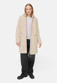Beige, fleecegefütterter Mantel mit Kapuze, Frontreißverschluss, zwei großen Taschen und lockerer Passform. Kombiniert mit einem leichten Oberteil und schwarzen Hosen.