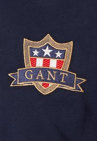 Tejido azul marino con un logotipo de escudo bordado en dorado, con franjas rojas y blancas, tres estrellas y una pancarta que dice "GANT".
