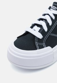 Melna audekla sporta kedu pāris ar baltām auklām. Iezīmējas ar biezu balto gumijas zoli un kontrastējošu baltu dūri gar augšdaļu. Apaļa purngalu forma.