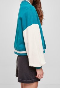 Kobieta w turkusowej i białej bomberce z długim rękawem, w czarnych szortach z podwiniętymi nogawkami, na jednolitym szarym tle.