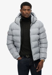 Giacca puffer grigia con cappuccio, caratterizzata da sezioni trapuntate, chiusura con zip e tasche laterali, abbinata a un maglione nero a colletto alto in maglia.