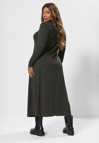 Frau mit langen, welligen Haaren, die ein dunkelgraues, langärmliges Maxikleid und schwarze Knöchelstiefel trägt, steht und schaut über ihre Schulter.