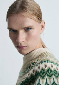 Vuna džemper s zelenim i krem uzorcima, s rebrastim ovratnikom ime mekanom teksturom. Krupni plan lica modela.