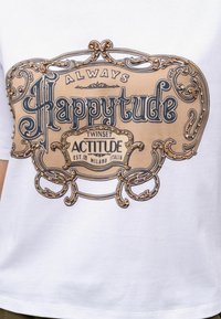 Maglietta bianca con una grafica centrale in beige e blu, caratterizzata da un lettering ornamentale e decorazioni, che recita "Always Happytude."