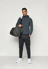 Grå zip-up jakke med hætte og sorte bukser, parret med hvide sneakers. Holder en sort sportsduffel med en stribet rem.