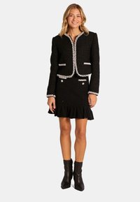 Ensemble en tweed noir comprenant une veste cropped avec un bord en perles et une jupe à volants, associée à des bottines noires.