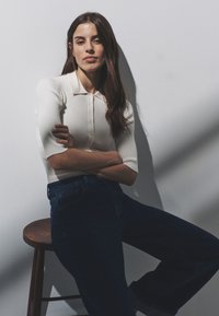 Vrouw met lang bruin haar, gekleed in een witte blouse met knopen en donkere spijkerbroek, zit op een houten kruk met gekruiste armen tegen een effen muur.