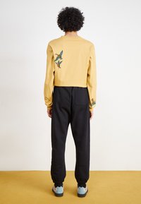 Yellow cropped long-sleeve top with embroidered birds on back, paired with black joggers. Socks and shoes feature blue accents. Textured fabric.