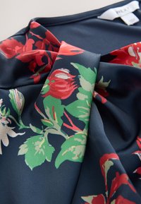 Haut en tissu bleu avec un motif floral rouge et vert, doté d'un nœud au niveau de l'encolure et d'une texture lisse et soyeuse.