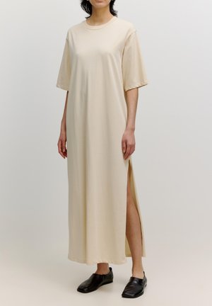 Beige maxi-jurk met korte mouwen en zijsplitten. Gemaakt van zachte stof met een relaxte pasvorm. Zwarte instappers completeren de outfit.