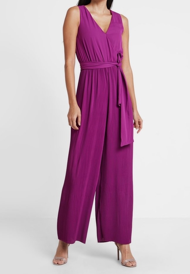 Femme portant une combinaison sans manches plissée violet profond avec jambes larges et une ceinture nouée à la taille, associée à des sandales à talons hauts couleur nude.