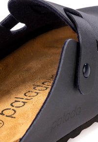 Zwarte sandaal met een gestructureerde bovenkant, tan suède binnenzool, opvallende stiksels en een knoopsluiting; minimalistisch ontwerp met uitsparingen.