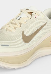 Κοντινή λήψη κρεμ και μπεζ παπουτσιού Nike για τρέξιμο, με υφασμάτινη επιφάνεια κυματιστής υφής, μπεζ λογότυπο Swoosh και παχιά μαξιλαρωτή σόλα με κόκκινη λεπτομέρεια.