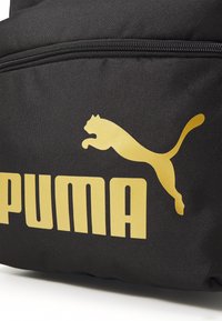 Fekete hátizsák, amelyen egy kiemelkedő arany Puma logó és márkanevének felirata található. Az anyag tartósnak tűnik, sima, textúrált felületű. A cipzáras zárás kiemelkedően látható.