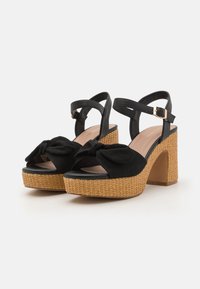 Sorte høyhælte sandaler med en vevd tan plattform og ankelreim, med en knyttet svart stoffdetalj foran.