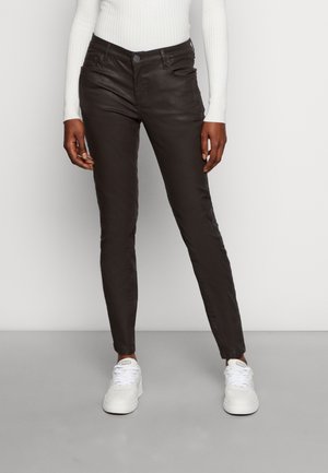 Persona che indossa pantaloni scuri aderenti, maglia bianca a maniche lunghe a costine e scarpe da ginnastica bianche in piedi davanti a uno sfondo semplice.