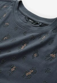 Tmavosivá tričko s krátkymi rukávmi, ktoré má textúrovaný vzor čiernych a metalických hnedých hrotov usporiadaných v tvare leopardieho vzoru.