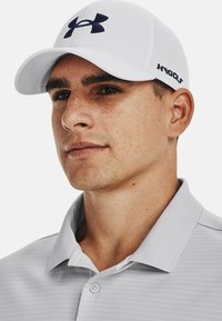 White baseball cap with dark blue Under Armour logo, paired with a light grey striped polo shirt. The fabric appears smooth and breathable.