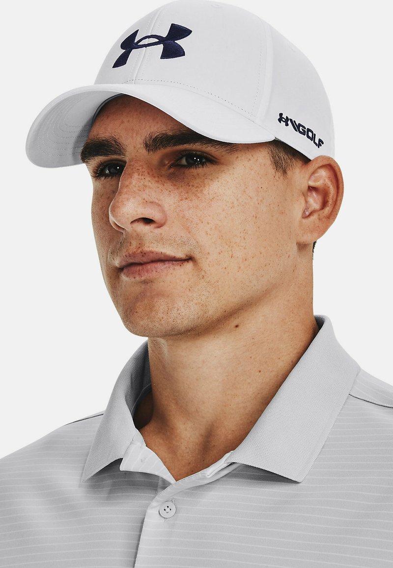 White baseball cap with dark blue Under Armour logo, paired with a light grey striped polo shirt. The fabric appears smooth and breathable.