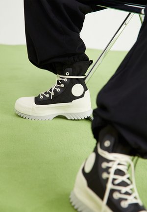Black and white high-top sneakers with thick ridged soles worn with black pants, resting on a light green floor under a metal chair.