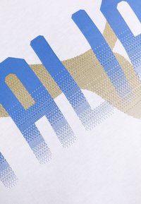 Camiseta blanca con un gran texto gráfico en azul y dorado, con un patrón de puntos en degradado. Material de algodón suave y textura lisa.