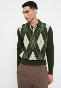 Grüner Poloshirt aus Strick mit Argyle-Muster in weißen und hellgrünen Rauten, mit Kragen und gerippten Bündchen. Kombiniert mit braunen, strukturierten Hosen.