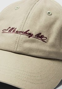 Cappellino da baseball in cotone beige con logo ricamato color marrone che recita "L'Ilot." Presenta una corona arrotondata e una fascia regolabile sul retro.
