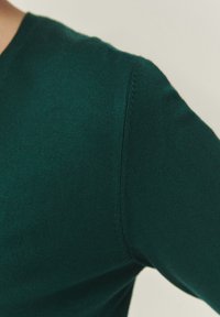 Pull vert foncé en maille avec un col en V, doté d'une texture lisse et d'un côtelé subtil aux coutures. Gros plan sur la zone de l'épaule.