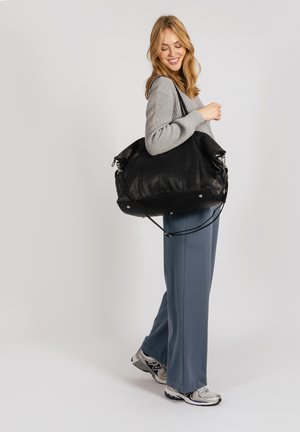 Black leather handbag with rounded shape, smooth texture, and silver hardware. Model wears grey sweater and blue pants, holding bag on shoulder.
