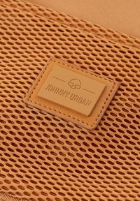 Gros plan d'un tissu en mesh marron avec un patch en cuir cousu portant l'inscription Johnny Urban et un petit logo au-dessus du texte.
