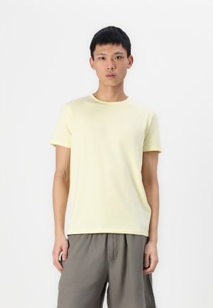 T-shirt en coton jaune clair avec un col rond et des manches courtes, avec un logo discret sur la poitrine gauche, associé à un short gris.