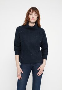 Pull en tricot côtelé bleu marine avec un col haut, orné de clous en argent décoratifs et d'une coupe décontractée. Manches longues et poignets côtelés.