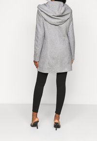 Mujer vestida con un abrigo gris con capucha, leggins negros y mule de tacón negro, de pie sobre un fondo blanco, de espaldas.