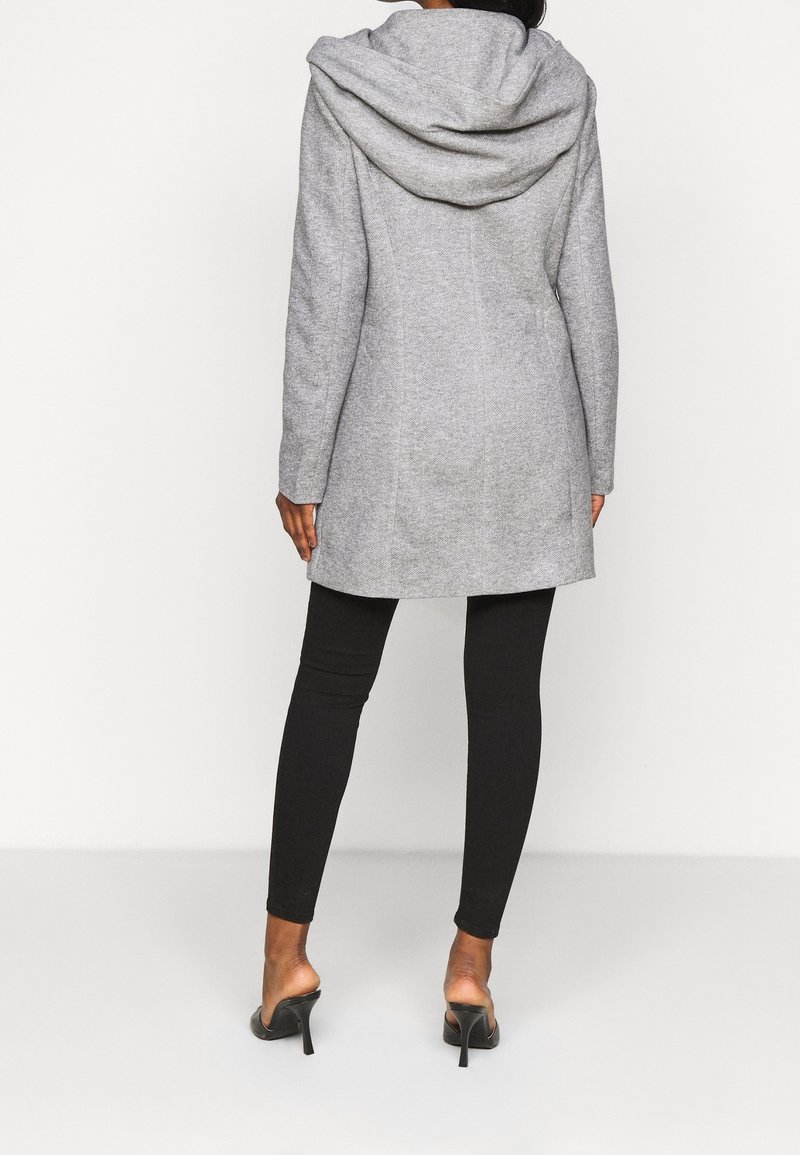 Mujer vestida con un abrigo gris con capucha, leggins negros y mule de tacón negro, de pie sobre un fondo blanco, de espaldas.