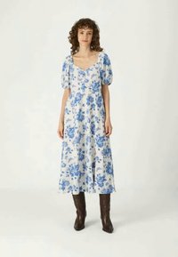 Bela cvetna obleka z modrimi rožnatimi vzorci, kratkimi naborki, kvadratnim izrezom in ohlapno silhueto, v kombinaciji s svetlo rjavimi gležnjarji.