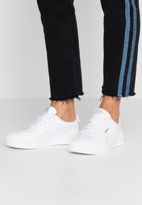 Weiße Sneakers mit strukturierten Gummisohlen, schlankem Design, Schnürverschluss und Markenakzenten. Kombiniert mit schwarzen Jeans mit blauen seitlichen Streifen.