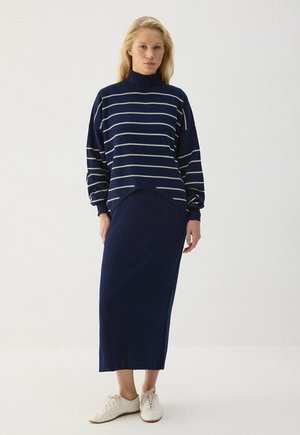 Femme portant un pull rayé bleu marine, une jupe côtelée assortie et des chaussures blanches décontractées, debout devant un fond clair uni.