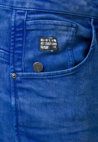 Blå denimjeans med en sidoficka med en silverknapp, G-Star-logga på midjebandet och ett strukturerat tygmönster.