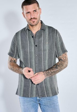 Mann mit kurzen Haaren und Tattoos trägt ein graues, gestreiftes Kurzarmhemd mit Knöpfen und hellblaue Jeans, er richtet sein Hemd mit beiden Händen.